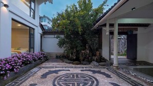 Um edifício moderno com área externa coberta, piso decorado com azulejos e um jardim com diversas plantas.