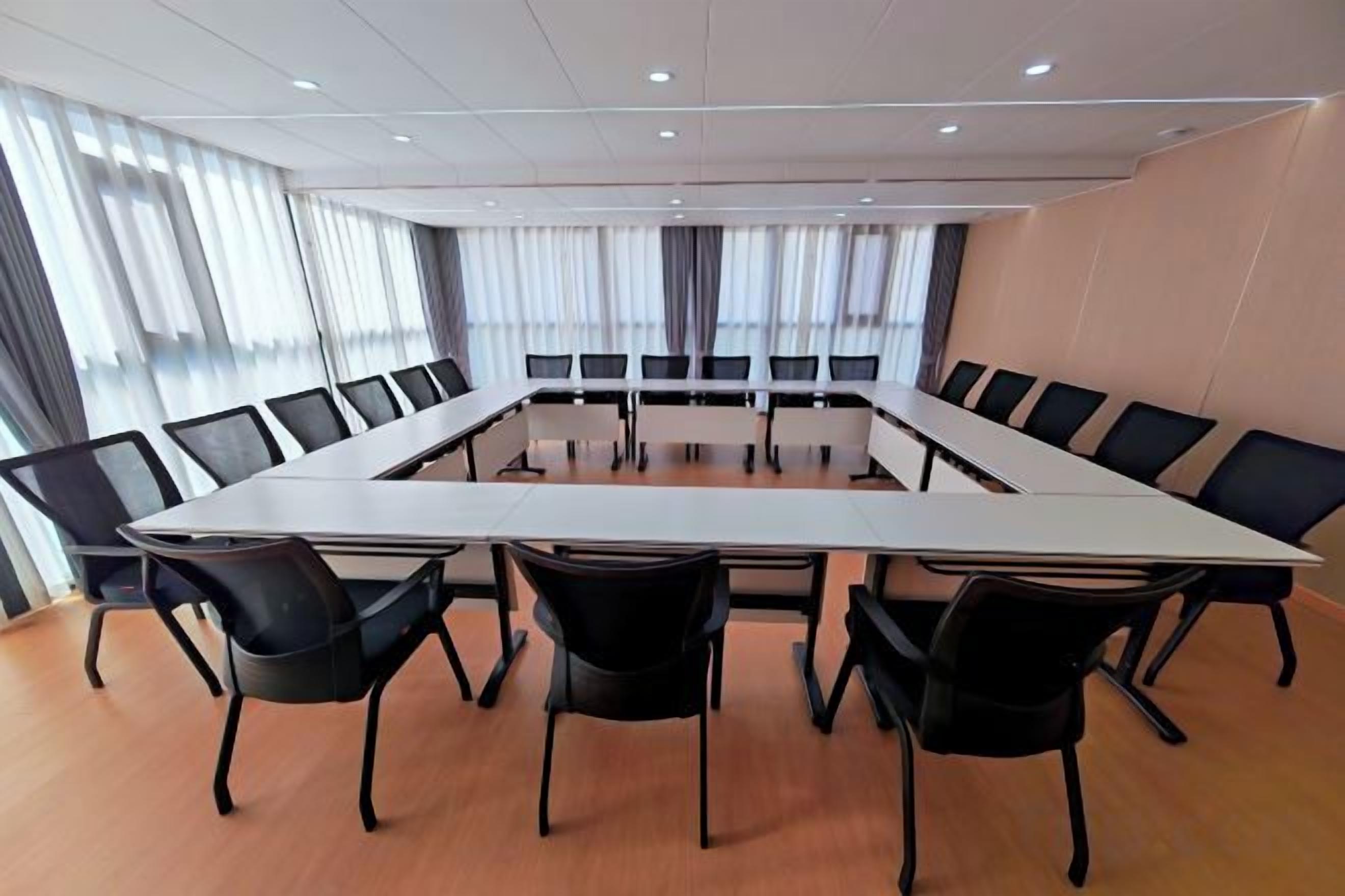 Een vergaderruimte met een lange tafel en stoelen, grote ramen met gordijnen, en een houten vloer.