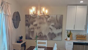 A dining area with a wooden table and white chairs, a chandelier, a painting on the wall, and a kitchen with white cabinets and a dishwasher. - Serene 3-bedroom waterfront newly renovated house, AC, Fast WiFi, Patio (White Marsh)