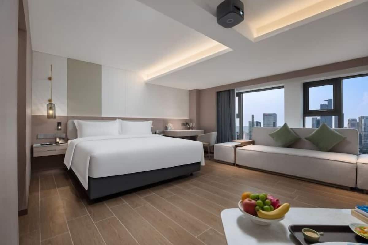 A modern hotel room with a large bed, a sofa, a dining table with fruit, and a city view.