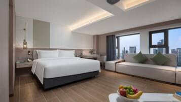 A modern hotel room with a large bed, a sofa, a dining table with fruit, and a city view.