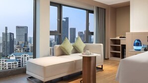 A modern hotel room with a large window offering a city view, a sofa with cushions, a bedside table, and a small desk with a lamp. - Sunflower Hotel & Residence Serviced apartment (Shenzhen)