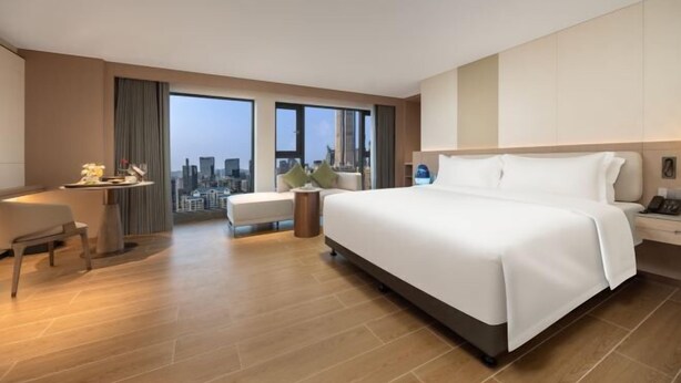 A modern hotel room with a large bed, a desk, a chair, a small table, and a view of the city. - Sunflower Hotel & Residence Serviced apartment (Shenzhen)