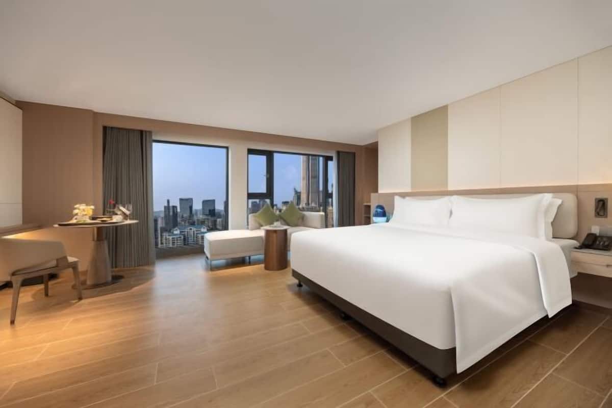 A modern hotel room with a large bed, a desk, a chair, a small table, and a view of the city.