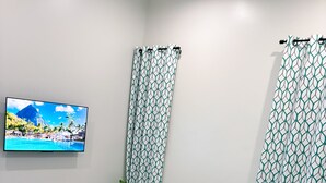 Un dormitorio con cama, ropa de cama color turquesa, almohadas blancas, un televisor en la pared y cortinas estampadas.