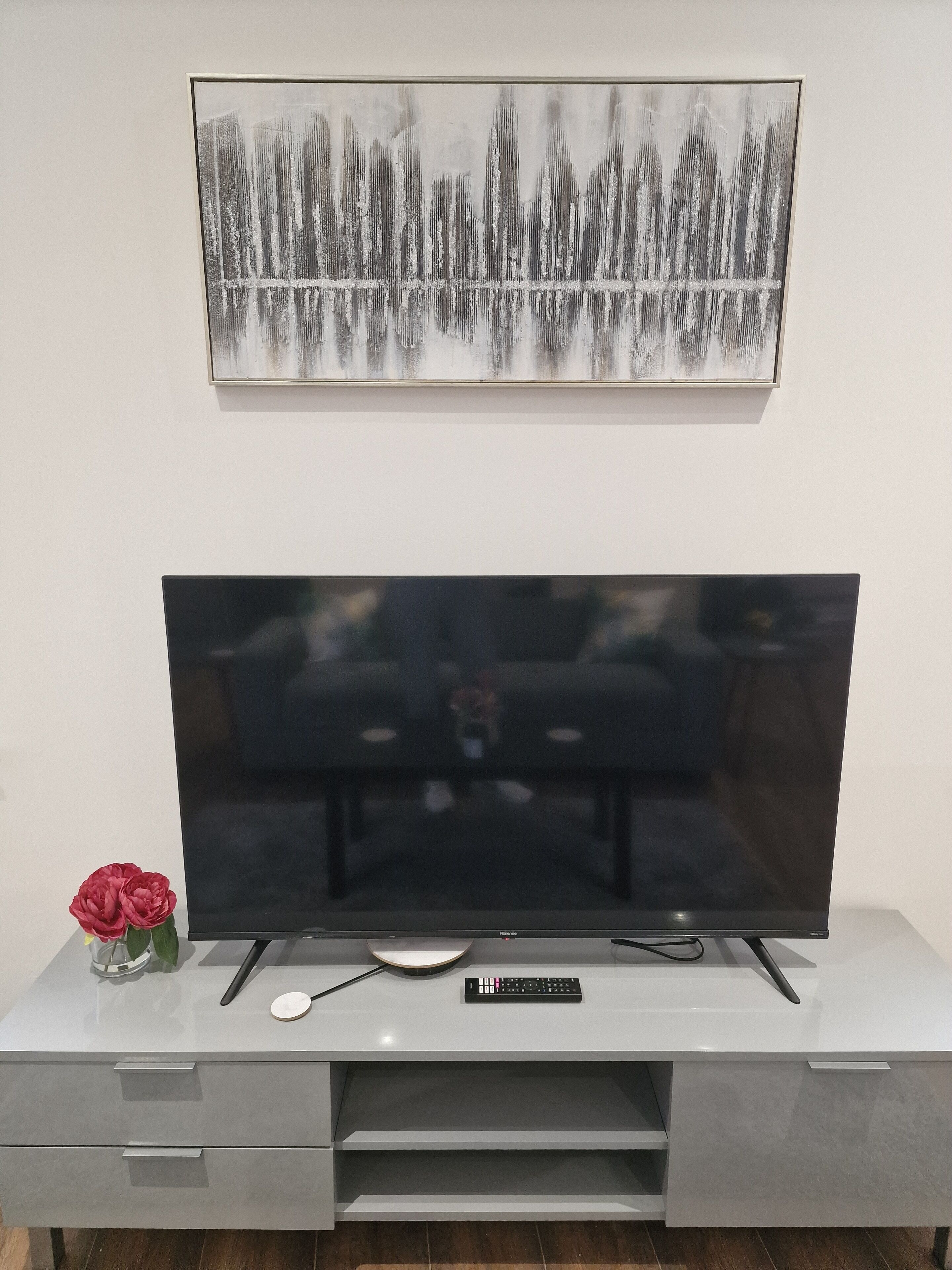 A modern living room with a flat-screen TV on a white entertainment unit, a framed abstract painting above the TV, and a vase with red flowers on the unit.