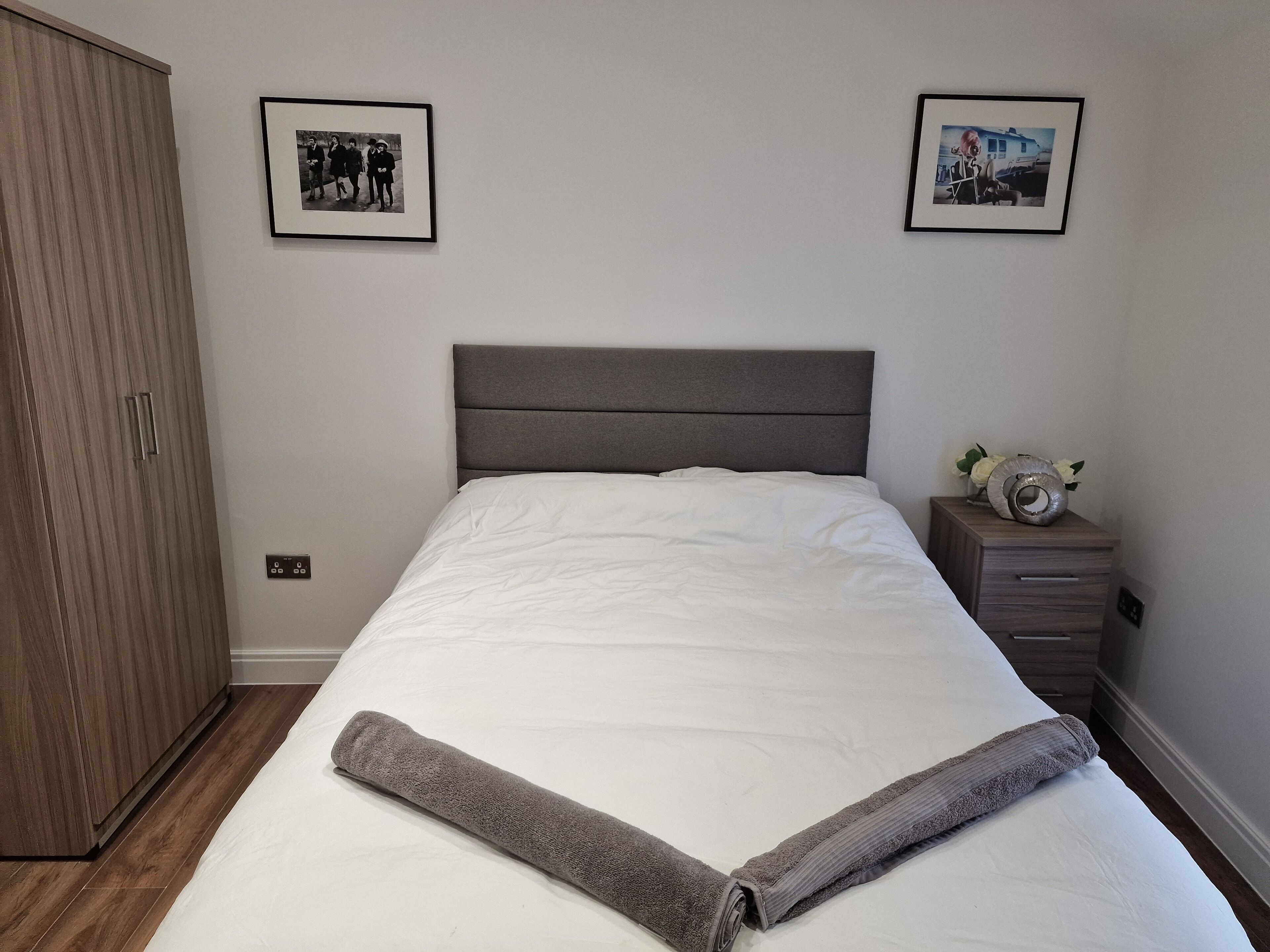 A neatly made bed with a grey headboard, a wooden wardrobe, and two framed pictures on the wall.