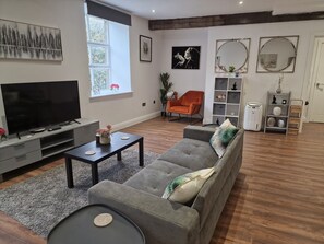 Uma sala de estar moderna com um sofá cinzento, uma mesa de centro, uma televisão sobre um móvel e uma janela com vista para árvores.
