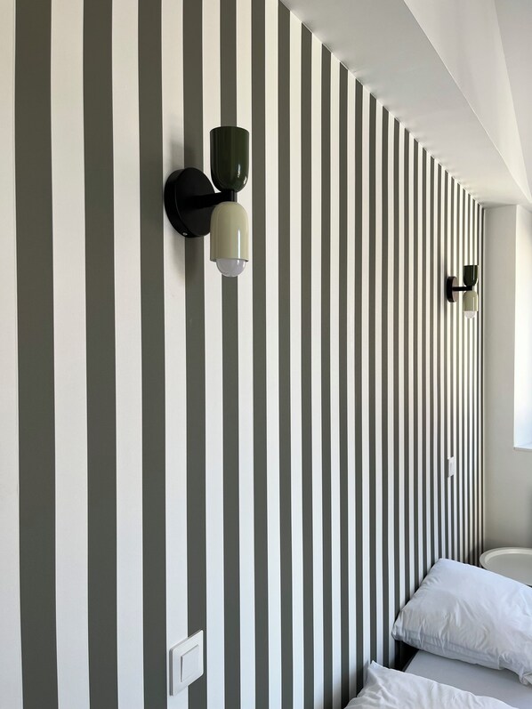 Una habitación con pared a rayas, dos lámparas fijadas a la pared, una cama con ropa blanca y una ventana.