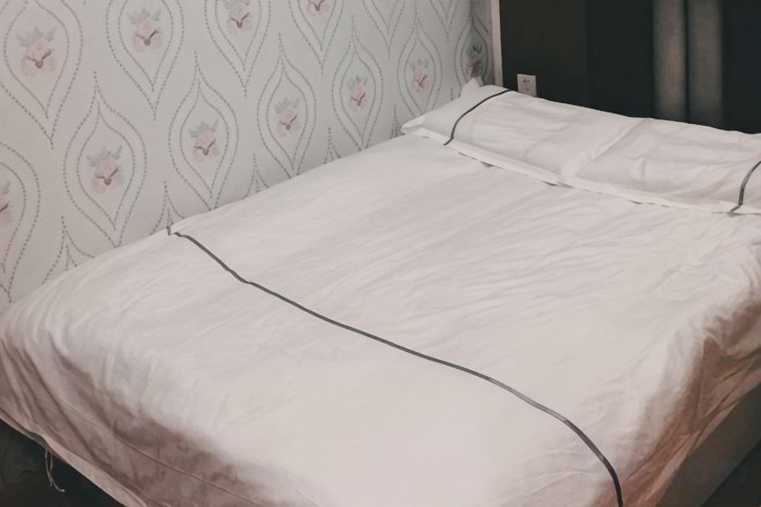 A bed with a white bedspread and matching pillows, a patterned wall behind it, and a visible power outlet on the wall.