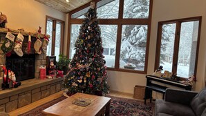 A cozy living room with a fireplace, a large Christmas tree, and a view of a snowy landscape through large windows. - 4-bedroom family home in charming Sun Valley close to Baldy and trails! (Sun Valley)