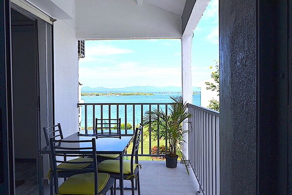 A balcony with a table and chairs, a potted plant, and a view of the sea and mountains.
