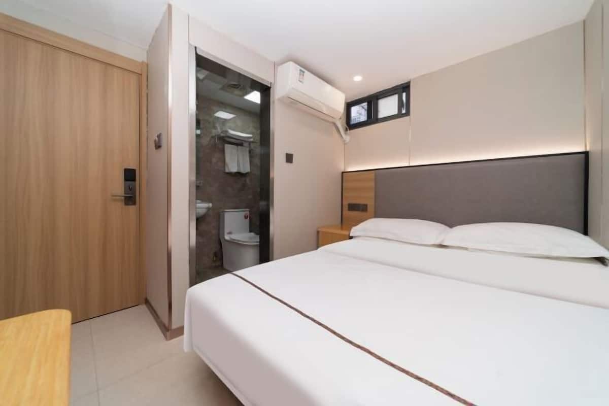 Una habitación de hotel moderna con una cama grande, un armario de madera, un baño con puerta de cristal y una mesita de noche pequeña.
