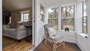Una sala de estar acogedora con un sofá gris, una mesita y un sillón de mimbre junto a un ventanal con vistas a árboles.