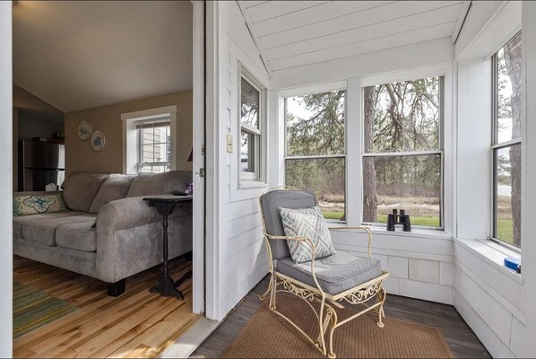 A cozy living room with a grey sofa, a small table, and a wicker chair by a large window with a view of trees.