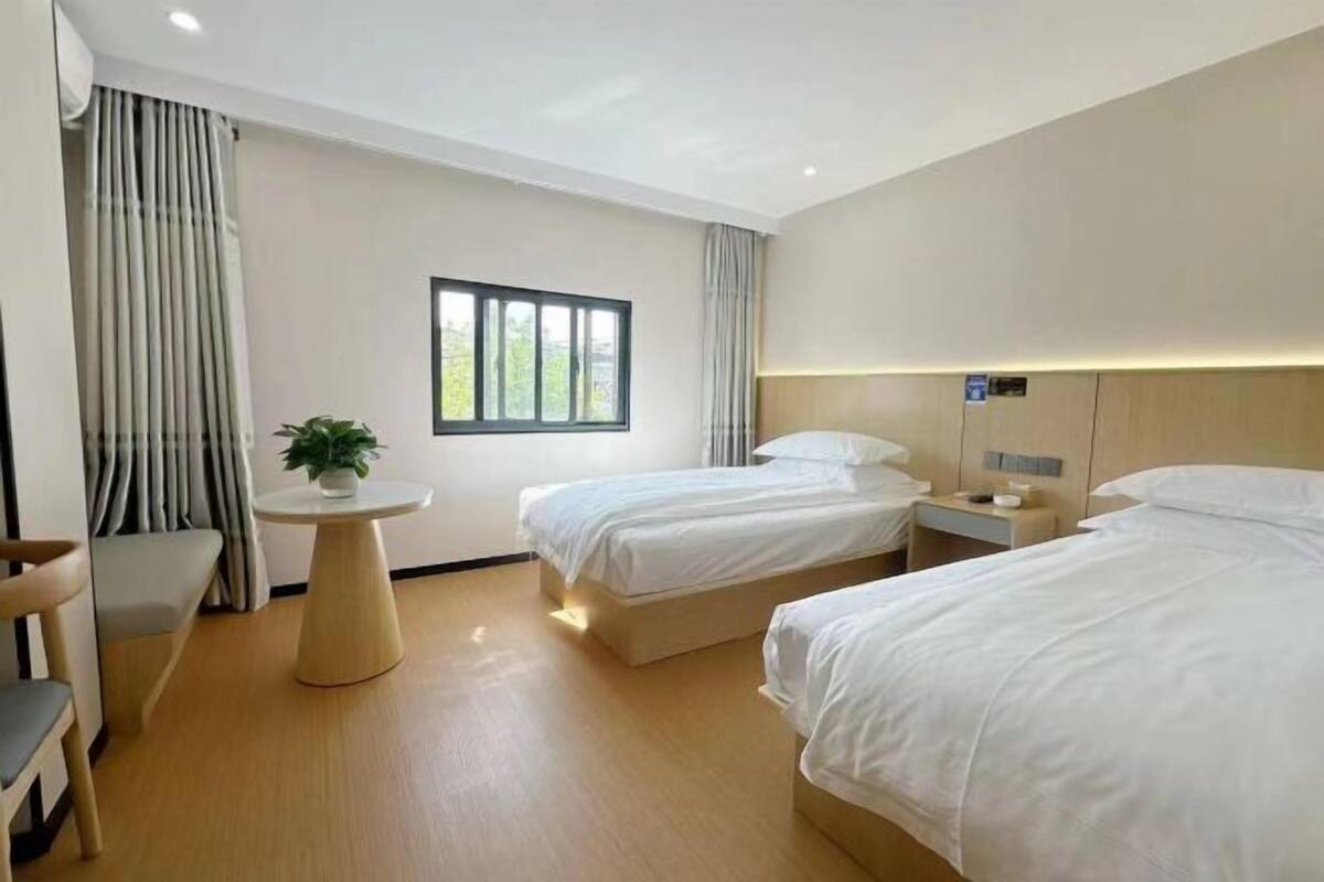 A hotel room with two beds, a small round table with a plant, a window, and a seating area with a chair.