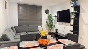 A modern living room with a grey sofa, a dining table with orange fruit, a potted plant, and a TV on the wall. - 1br-sleeps2-parking-nearlondon-central (Erith)