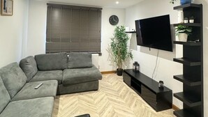 A modern living room with a sectional sofa, a flat-screen TV mounted on the wall, a black entertainment unit, and a potted plant. - 1br-sleeps2-parking-nearlondon-central (Erith)