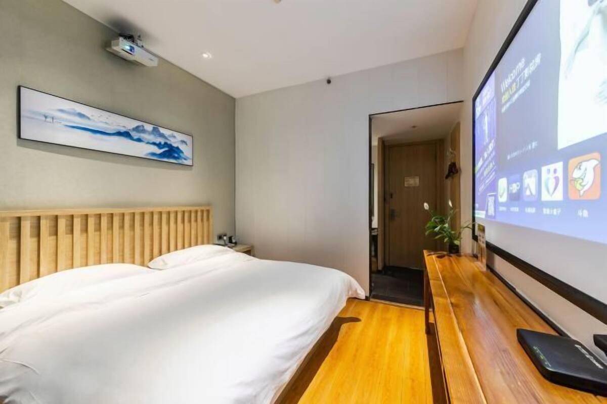 Une chambre d’hôtel avec un lit, une tête de lit en bois, un écran de projection et un banc en bois.
