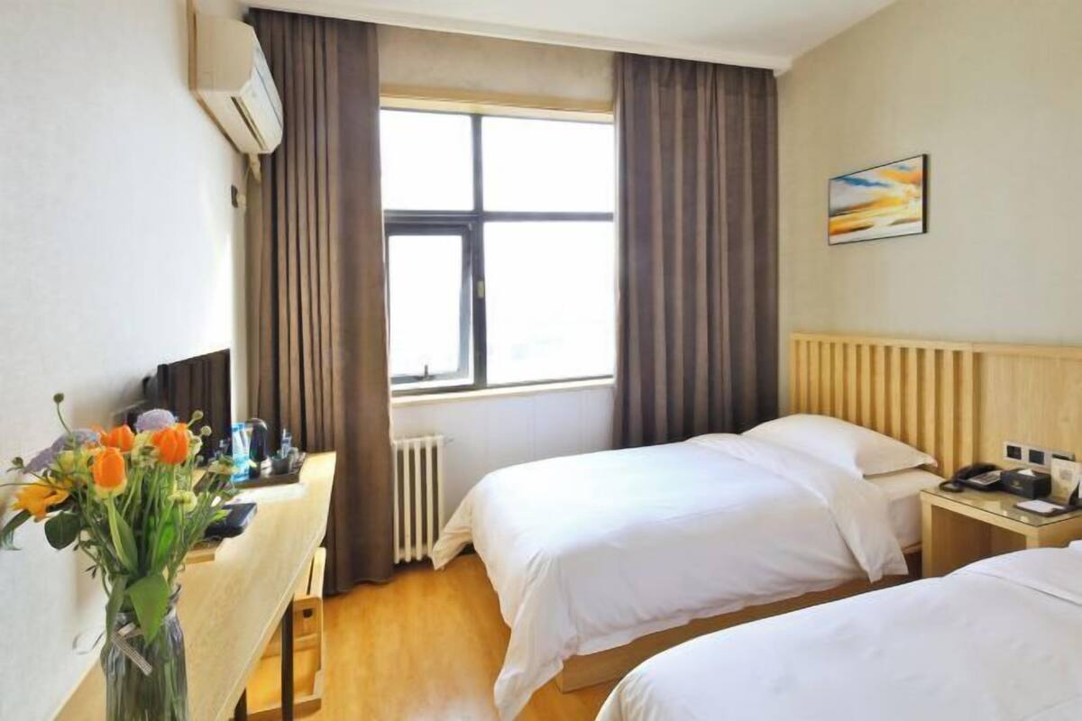 Une chambre d’hôtel avec deux lits, un bureau agrémenté d’un vase de fleurs, un radiateur, une fenêtre avec des rideaux et un tableau accroché au mur.