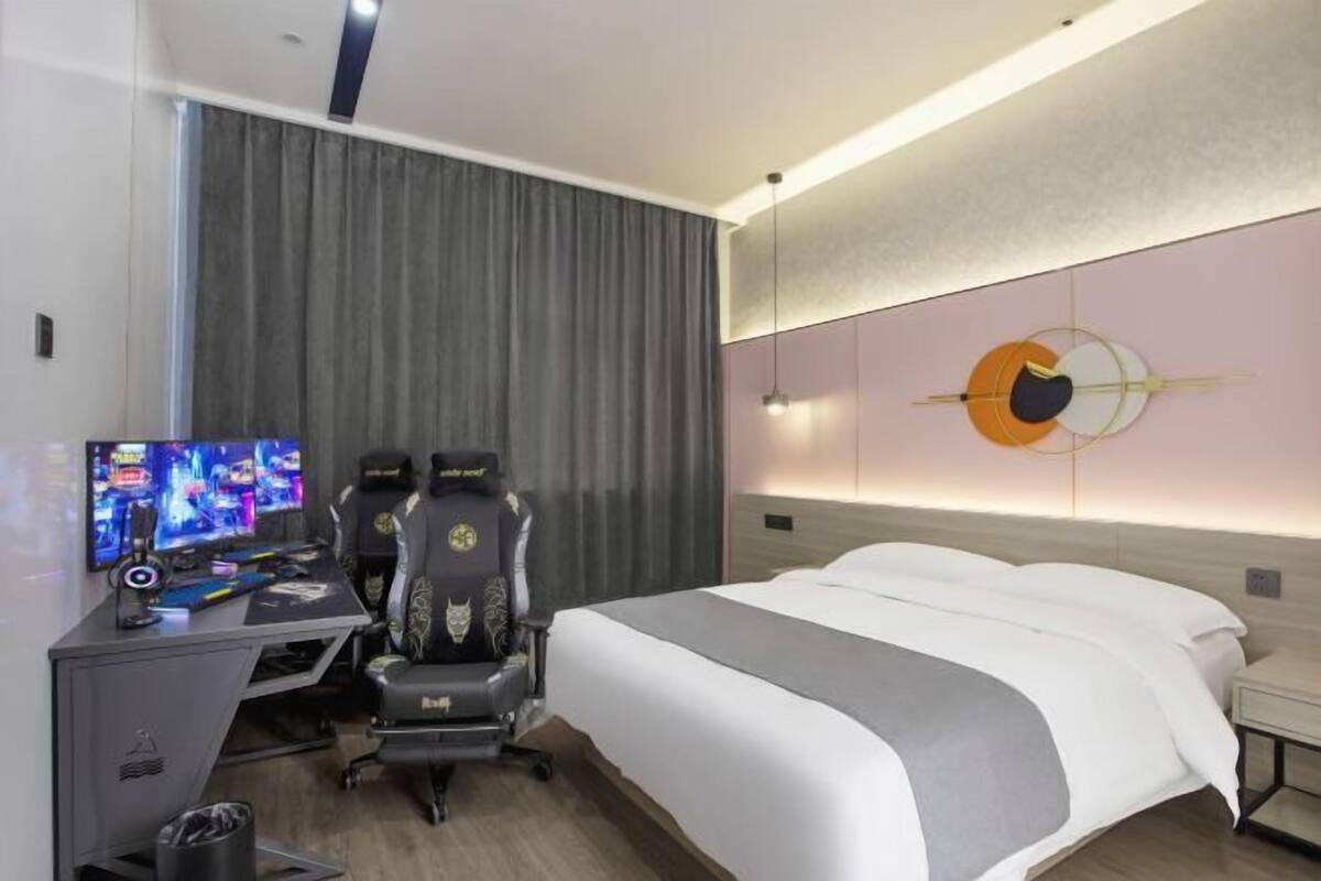 Une chambre d’hôtel comprenant un lit, un bureau avec un ordinateur et un fauteuil de jeu, ainsi qu’une œuvre d’art accrochée au mur.