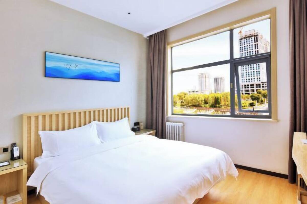 Une chambre d’hôtel avec un grand lit, une table de chevet, un tableau et une fenêtre donnant sur des immeubles et des arbres.