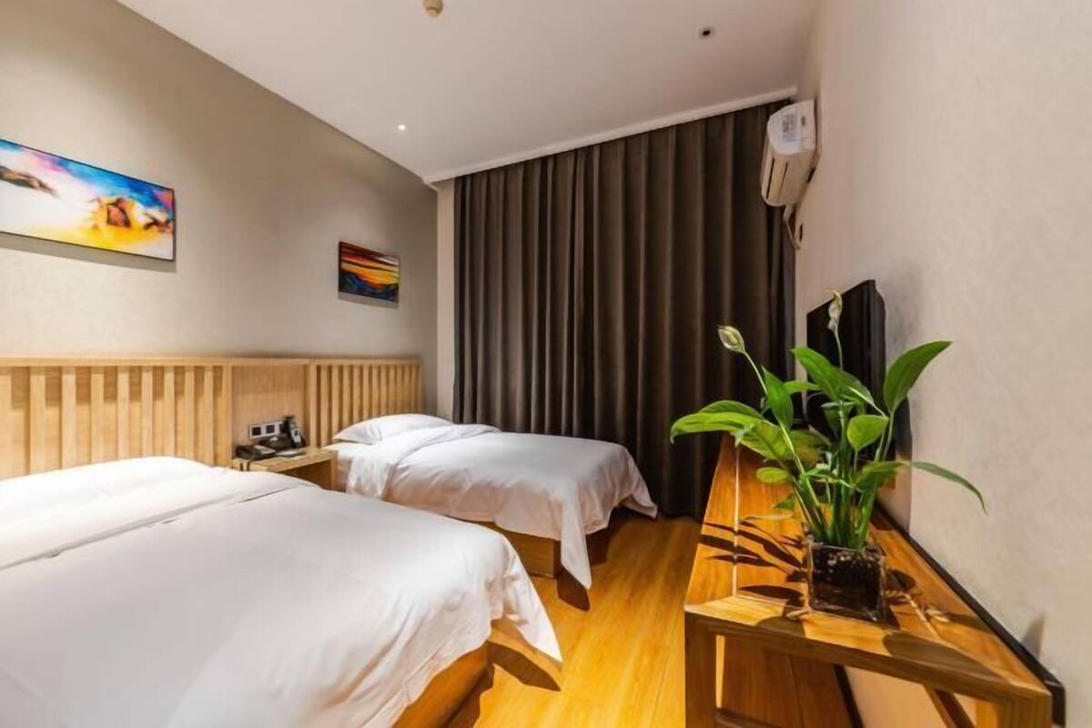 Une chambre d’hôtel avec deux lits, une tête de lit en bois, une table de chevet avec une plante, un tableau au mur et une grande fenêtre avec des rideaux.