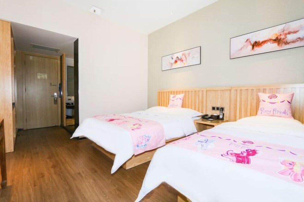Een hotelkamer met twee bedden, elk met een roze deken met tekenfilmfiguren. Twee ingelijste foto’s aan de muur, een houten deur en een houten vloer.