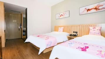 A hotel room with two beds, each with a pink blanket featuring cartoon characters. There are two framed pictures on the wall, a wooden door, and a wooden floor.