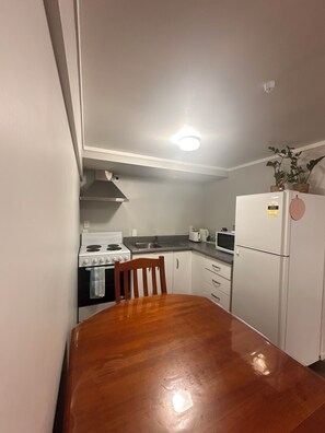 A compact kitchen with white cabinets, a stove, microwave, and a refrigerator. There is a wooden dining table and a chair. - A cosy, private and convenient space. 8-minute walk to Rotorua Stadium! (Rotorua)