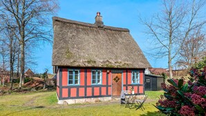 Une maison traditionnelle à colombages, avec un toit de chaume, un petit porche et un jardin agrémenté d’une chaise et d’une table.