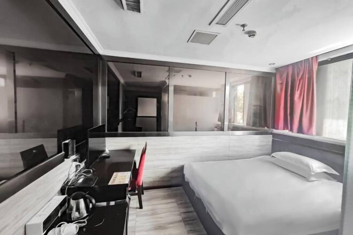 Une chambre d’hôtel moderne équipée d’un lit, d’un bureau avec une chaise, d’un grand miroir et d’une fenêtre avec des rideaux.
