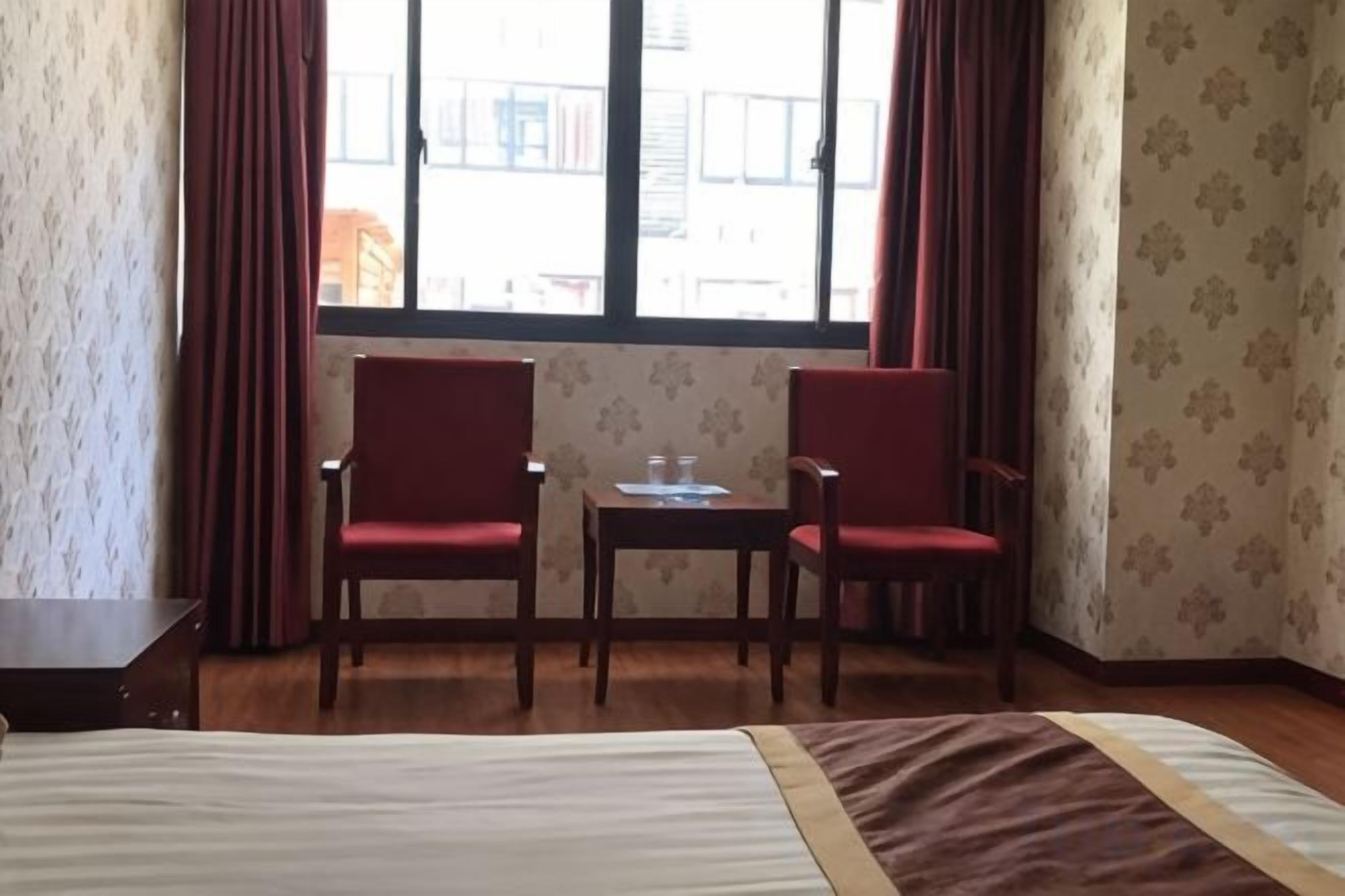 Quarto de hotel com cama, duas cadeiras vermelhas, uma mesinha e janela com cortinas.
