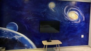 A room with a large mural of a galaxy on the wall, a flat-screen TV mounted above a wooden bench, and a metal bowl on the floor. - FanYing Business Hotel (Huzhou)
