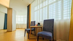 A hotel room with a wooden floor, a small round table with a coffee maker, two blue chairs, and large windows with sheer curtains. - Anxin Intercity Hotel (Quanzhou Quangang) (Quanzhou)