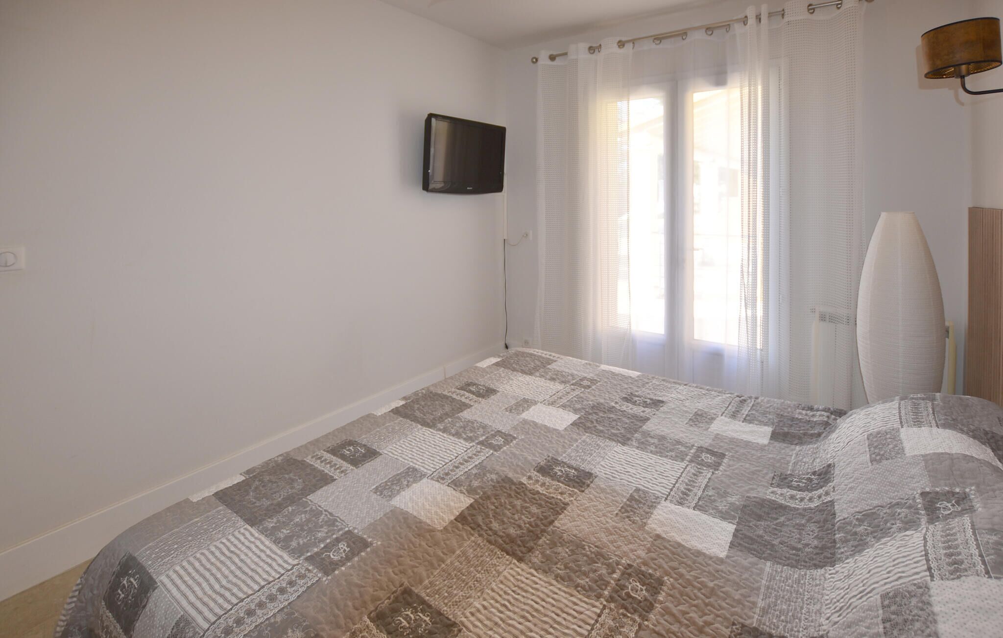 Een slaapkamer met een groot bed, een flatscreen televisie aan de wand en een raam met dunne gordijnen.