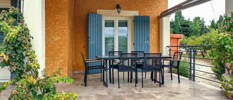 An outdoor patio with a table and chairs, a door with blue shutters, and a stone-tiled floor.