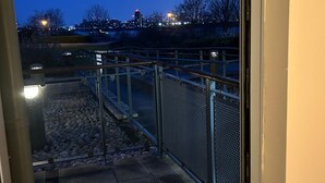 A view through a glass door of a parking area with a metal fence, a paved walkway, and a city skyline in the background. - Flat in Greenwich Near O2 and London City Airport (London)