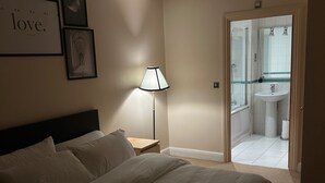 A bedroom with a bed, a nightstand, a lamp, and a door leading to a bathroom. - Flat in Greenwich Near O2 and London City Airport (London)