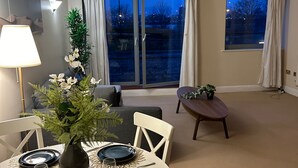 A dining area with a round table set for four, a vase with flowers, and a view of the outdoors through large windows. - Flat in Greenwich Near O2 and London City Airport (London)