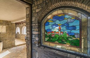 Stained glass window features a church and landscape scene, set within a stone wall.