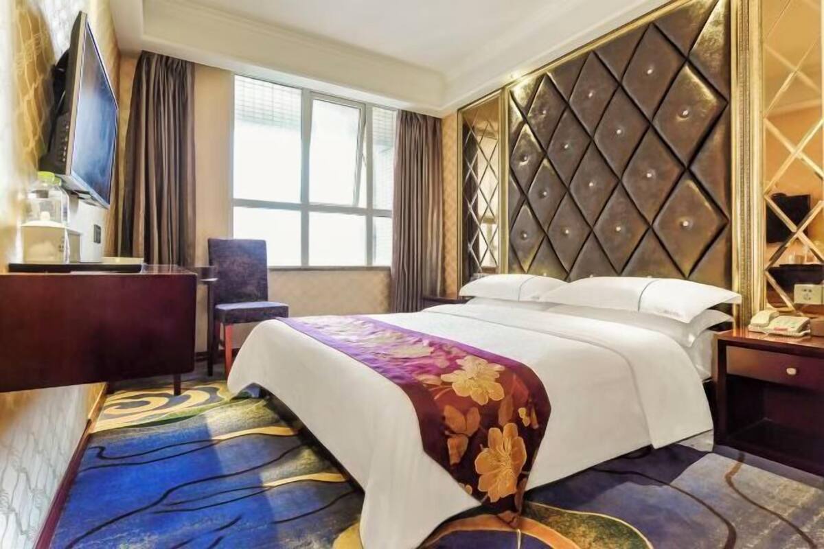 A hotel room with a large bed, a TV, a desk, and a window with curtains.