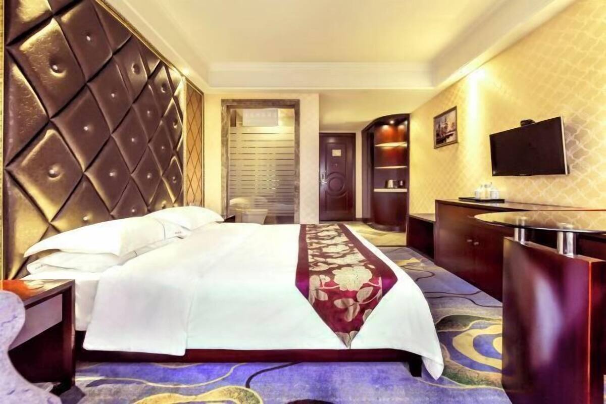 A hotel room with a tufted headboard, a large bed with a white and red bedspread, a flat-screen TV mounted on the wall, and a small desk with a mirror and a lamp.