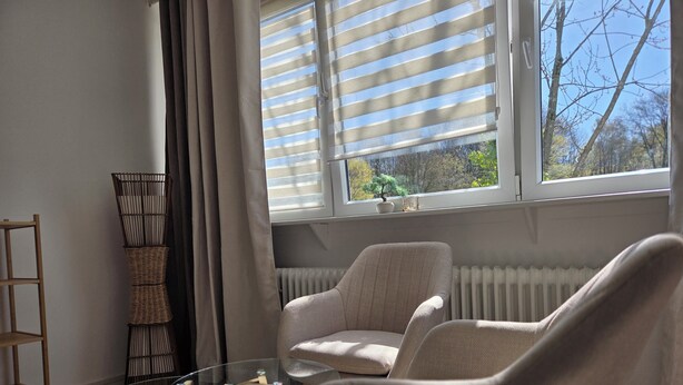 Ett rum med ett stort fönster, ett element, en flätad stol, ett glasbord och en hylla i trä.