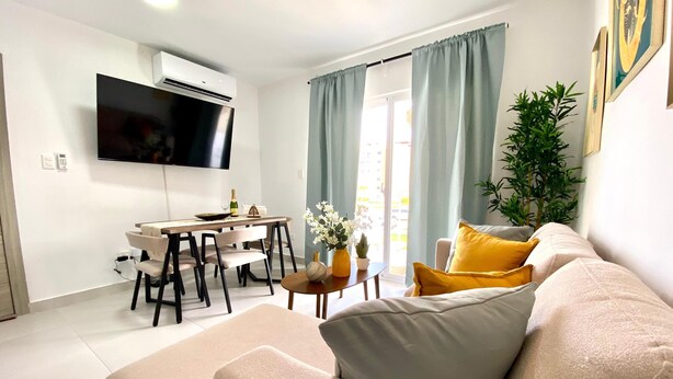 Una sala de estar moderna con un sofá color beige, un comedor con mesa y sillas, un televisor de pantalla plana en la pared y un amplio ventanal con cortinas.