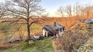 A rustic house with a sloped roof, surrounded by a grassy landscape and trees.