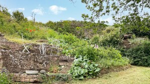 A rustic outdoor seating area with metal chairs and a table, surrounded by lush greenery and a stone wall.
