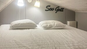 A neatly made bed with two pillows and a quilted blanket, a wall-mounted light fixture, and a wall sign that reads "Sow Gott".