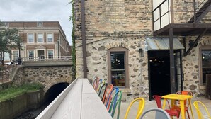 A narrow water channel with a wooden walkway, stone buildings, and outdoor seating with colorful chairs.