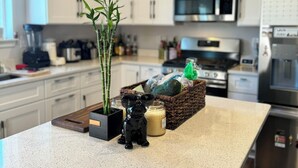 A modern kitchen with white cabinets, stainless steel appliances, a central island with a plant, a decorative dog figurine, and a wicker basket with groceries.